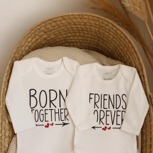 Könnte beinhalten: Zwei weiße Baby-Bodys mit langen Ärmeln, jeweils mit schwarzem Text. Einer lautet "BORN TOGETHER" mit Pfeilen und Herzen, der andere "FRIENDS FOREVER" mit Pfeilen und Herzen. Sie liegen in einem Weidenkorb.