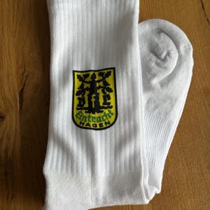 May include: White ribbed socks with a black, yellow, and green emblem that reads "Eintracht Hagen". The socks are folded on a wooden surface. The emblem features a tree design.