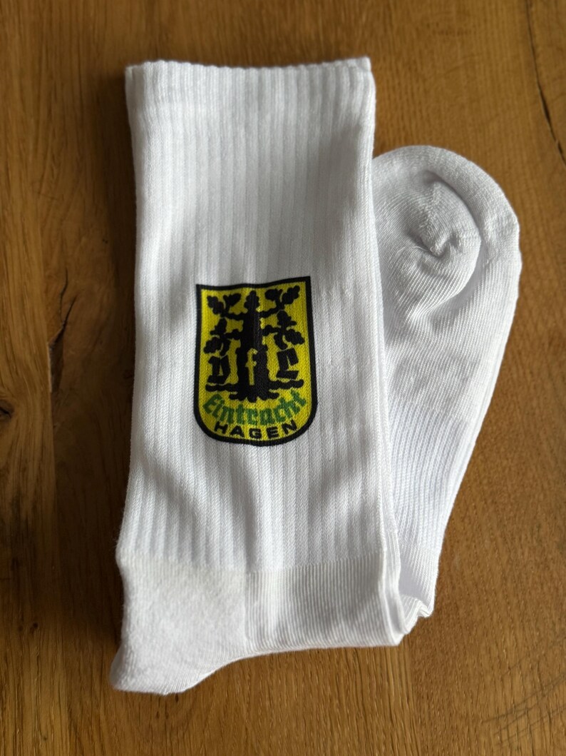 May include: White ribbed socks with a black, yellow, and green emblem that reads "Eintracht Hagen". The socks are folded on a wooden surface. The emblem features a tree design.