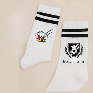 May include: Pair of white athletic socks with black stripes at the top. One sock features a colorful emblem with text, while the other displays a soccer-themed crest and the words "Bester Trainer."