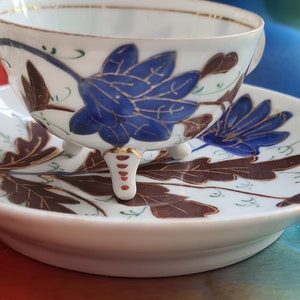 Pode incluir: Uma chávena de chá e um pires de porcelana branca com um desenho floral. A chávena tem três pés dourados e o pires tem um padrão floral castanho e azul.