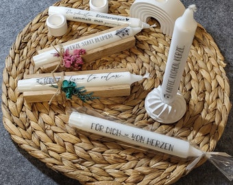 May include: Assortment of white candles with various text printed on them, including "Dahon Erinnerungen sammelt man gerne", "KERZE STATT BLUMEN", "time to relax", "FÜR DICH VON HERZEN", and "KAFFEE, KUCHEN, KERZE". The candles are displayed on wooden bases and a white ceramic base.