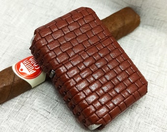 May include: A brown leather cigar case with a woven pattern is open, revealing a cigar. The cigar has a white and red label. The case and cigar rest on a white, textured surface.