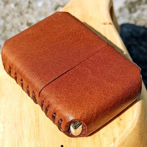 May include: A brown leather case with a stitched edge and a metal accent. The case is rectangular with rounded corners and a textured surface. The leather has a rich, warm tone.
