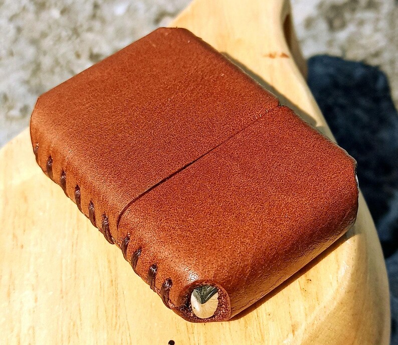 May include: A brown leather case with a stitched edge and a metal accent. The case is rectangular with rounded corners and a textured surface. The leather has a rich, warm tone.