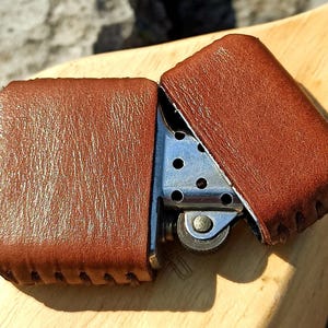May include: A brown leather lighter case, open to reveal the silver lighter inside. The leather case has a textured surface and is stitched along the edges. The lighter is on a wooden surface.