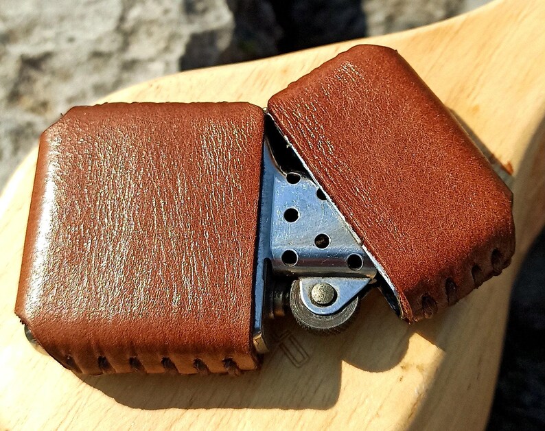May include: A brown leather lighter case, open to reveal the silver lighter inside. The leather case has a textured surface and is stitched along the edges. The lighter is on a wooden surface.