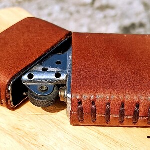 May include: A brown leather lighter case with a silver lighter inside. The case is open, revealing the lighter's metal components. The leather has visible stitching and a textured surface.