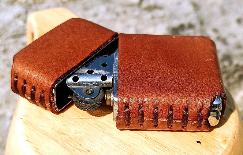May include: A brown leather lighter case with a silver lighter inside. The case is open, revealing the lighter's metal components. The leather has visible stitching and a textured surface.