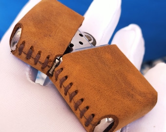 May include: A brown leather lighter case with stitching detail. The case is closed and has a silver metal lighter inside.