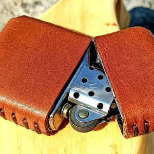 May include: A brown leather lighter case, open to reveal a silver metal lighter. The case is made of textured brown leather with stitched edges. The lighter has a wheel and a small opening for the flame.