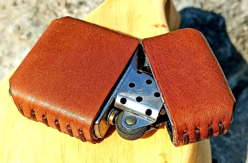 May include: A brown leather lighter case, open to reveal a silver metal lighter. The case is made of textured brown leather with stitched edges. The lighter has a wheel and a small opening for the flame.