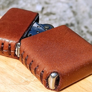 May include: A brown leather lighter case with a stitched design. The case is open, revealing the lighter inside. The leather has a textured appearance and a metal accent on the bottom.