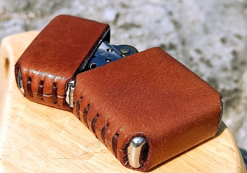 May include: A brown leather lighter case with a stitched design. The case is open, revealing the lighter inside. The leather has a textured appearance and a metal accent on the bottom.