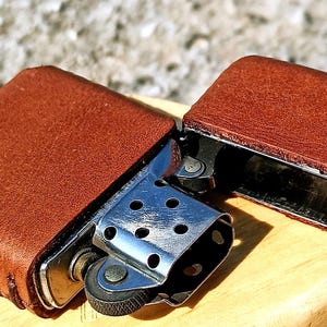 May include: A brown leather lighter case, open to reveal the silver lighter mechanism. The case is made of textured brown leather and is stitched along the edges. The lighter is resting on a wooden surface.