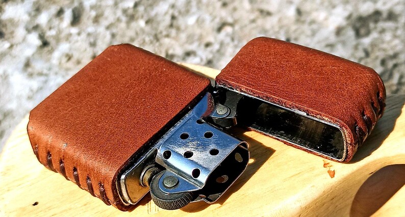 May include: A brown leather lighter case, open to reveal the silver lighter mechanism. The case is made of textured brown leather and is stitched along the edges. The lighter is resting on a wooden surface.