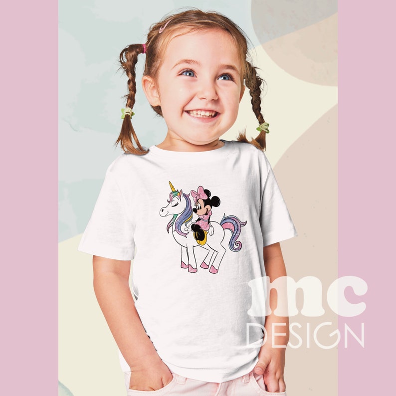 May include: White t-shirt featuring a cartoon illustration of a unicorn with a pink and purple mane and tail, ridden by a character wearing a pink dress and bow. The shirt has the text "mc DESIGN" in the lower right corner.