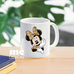 May include: White ceramic mug with a cartoon image of Minnie Mouse wearing a yellow and black plaid bow and dress. The mug has a black handle.