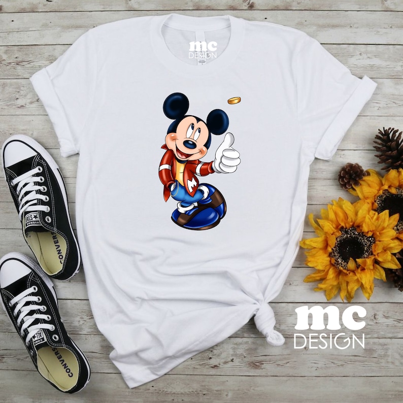 K&ouml;nnte beinhalten: Wei&szlig;es T-Shirt mit einem Cartoon-Bild von Mickey Mouse, der eine rote Jacke und eine blaue Jeans tr&auml;gt. Mickey Mouse zeigt den Daumen nach oben und hat eine goldene M&uuml;nze &uuml;ber seinem Kopf. Der Text "mc DESIGN" ist auf das T-Shirt gedruckt.
