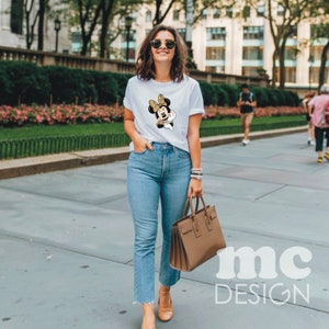 May include: A woman wearing a white t-shirt with a cartoon image of Minnie Mouse. She is wearing blue jeans and brown shoes. She is carrying a brown handbag.