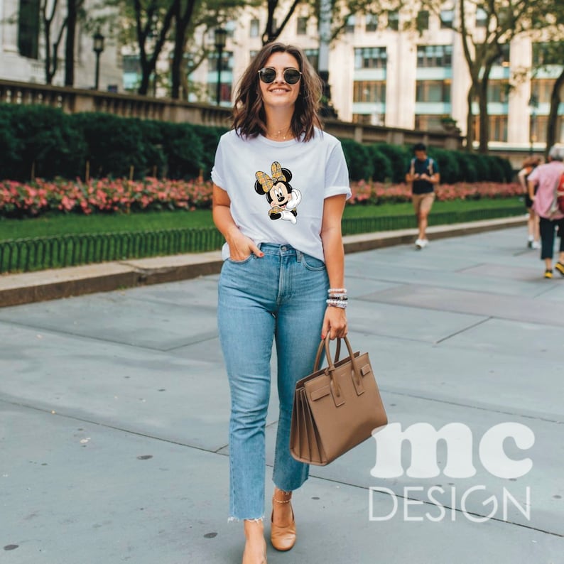 May include: A woman wearing a white t-shirt with a cartoon image of Minnie Mouse. She is wearing blue jeans and brown shoes. She is carrying a brown handbag.