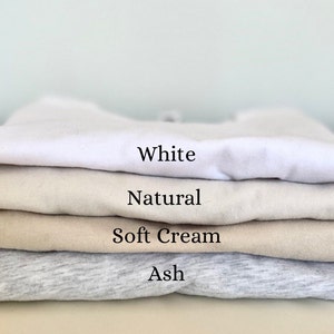 May include: A stack of four folded t-shirts in different colors: white, natural, soft cream, and ash.