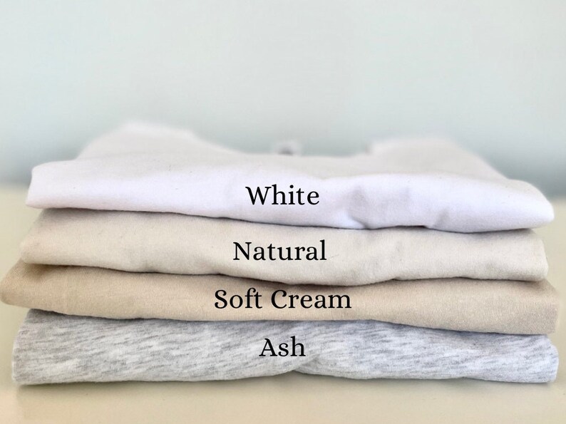 May include: A stack of four folded t-shirts in different colors: white, natural, soft cream, and ash.