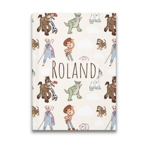 May include: A rectangular notebook with a light beige cover featuring a repeating pattern of cartoon characters including a cowboy, a cowgirl on a horse, a green dinosaur, a shepherdess, a dachshund, and a pig. The name ROLAND is printed in brown.