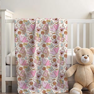 Peut inclure: Une couverture de b&eacute;b&eacute; blanche avec un motif sur le th&egrave;me de Mickey Mouse en rose et marron. Le motif comprend des oreilles de Mickey Mouse, des seaux de pop-corn, des cornets de glace et des cupcakes.