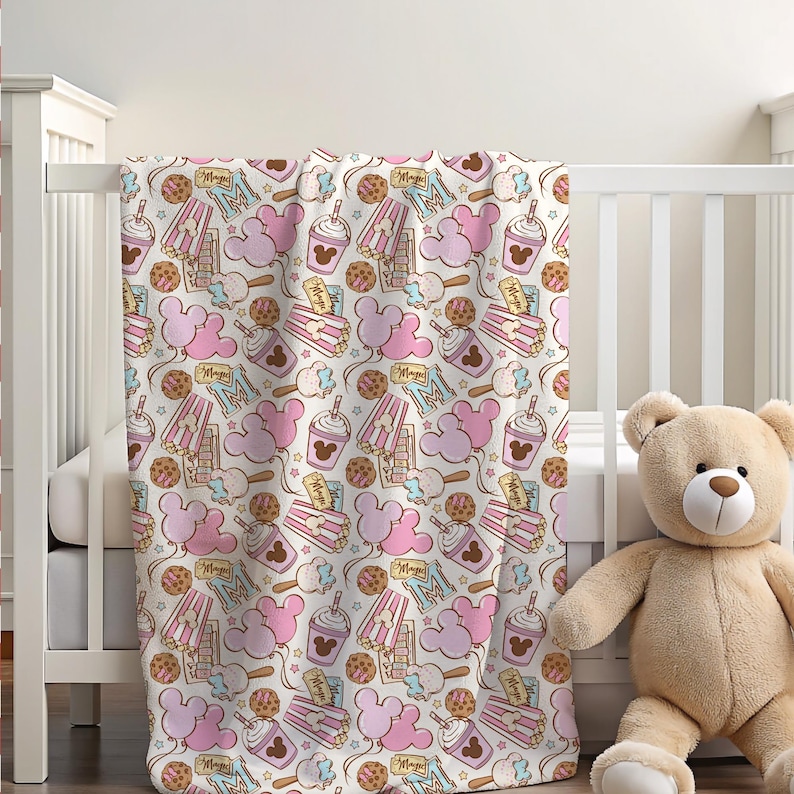 Peut inclure: Une couverture de b&eacute;b&eacute; blanche avec un motif sur le th&egrave;me de Mickey Mouse en rose et marron. Le motif comprend des oreilles de Mickey Mouse, des seaux de pop-corn, des cornets de glace et des cupcakes.