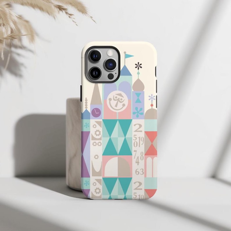 Puede incluir: Una funda blanca para iPhone con un dise&ntilde;o geom&eacute;trico colorido que presenta un castillo caprichoso con un reloj, n&uacute;meros y una cara sonriente. La funda tiene un recorte para el objetivo de la c&aacute;mara.