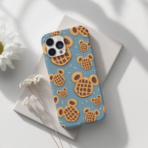 May include: A blue phone case featuring a repeating pattern of waffle-shaped designs with Mickey Mouse ears. The waffles are brown with a grid pattern, and the background has small white dots. The case has a camera cutout.