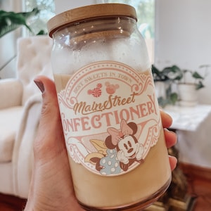 May include: A glass jar with a bamboo lid filled with a light brown liquid. The jar has a pink and white sticker with the text "Main Street Confectionery" and a cartoon image of Minnie Mouse.