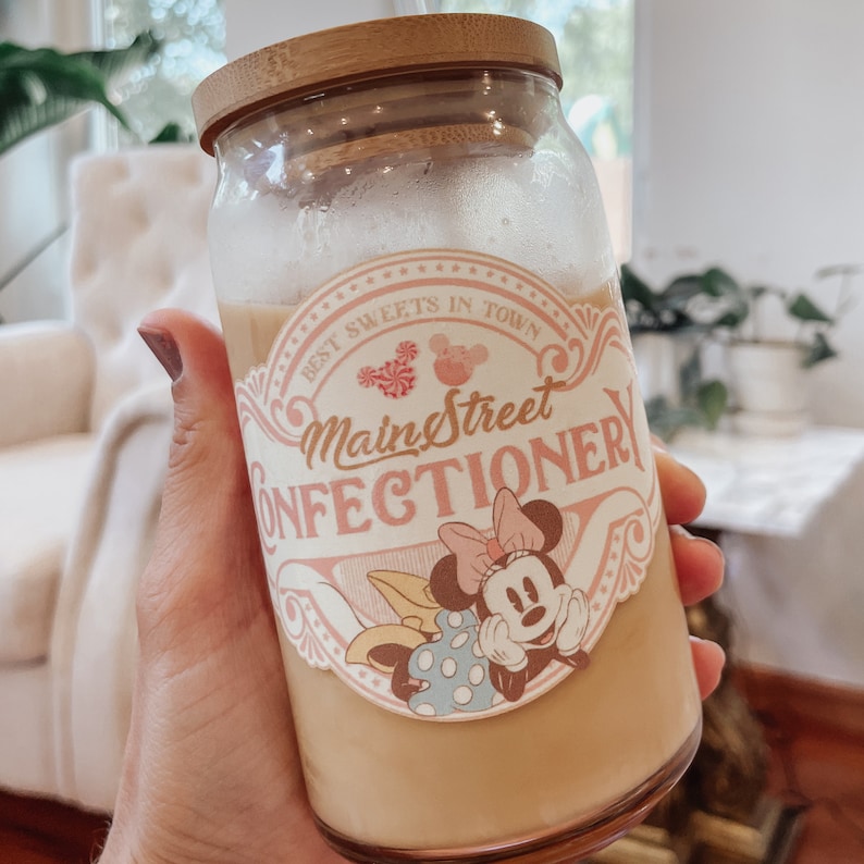 May include: A glass jar with a bamboo lid filled with a light brown liquid. The jar has a pink and white sticker with the text "Main Street Confectionery" and a cartoon image of Minnie Mouse.