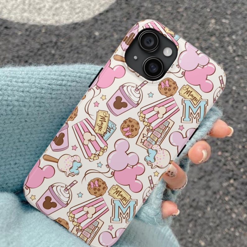 May include: A white smartphone case featuring a repeating pattern of pink mouse-shaped balloons, popcorn buckets, chocolate chip cookies, and cups with straws. The design includes light blue text that reads Magic and the letter M.