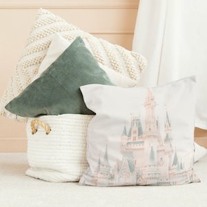 Peut inclure: Un coussin blanc avec une image rose et blanche du château de Cendrillon. Le coussin est posé sur un panier blanc en osier avec deux autres coussins, un vert et un blanc.