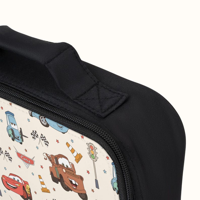 May include: Black lunch bag with a zipper closure and a top handle. The bag is decorated with a white background and a repeating pattern of cartoon cars, stars, and checkered flags. The text "Cars" is visible on the bag.