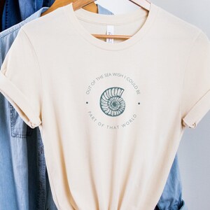 May include: A beige t-shirt with a graphic of a seashell and the text "Out of the sea wish I could be part of that world."