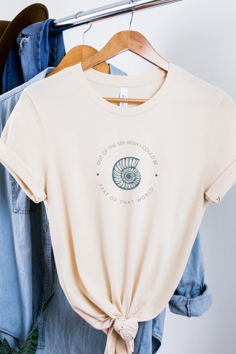 May include: A beige t-shirt with a graphic of a seashell and the text "Out of the sea wish I could be part of that world."