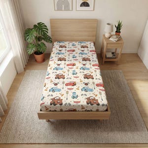 May include: A twin-sized bed featuring a fitted sheet with a repeating pattern of cartoon race cars, traffic cones, and checkered flags on a cream background. The bed has a light wood frame and a matching headboard, positioned in a room setting.