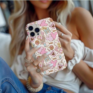 May include: A smartphone case with a cream background featuring a repeating pattern of pink mouse-shaped balloons, popcorn boxes, chocolate chip cookies, and drinks. The design includes the letter M and the word Magic in gold-toned text.