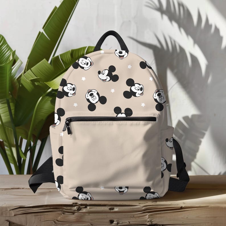 Puede incluir: Una mochila beige con cremallera y correas negras. La mochila tiene un patr&oacute;n repetitivo de cabezas de Mickey Mouse con estrellas blancas.