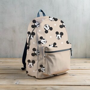 Puede incluir: Una mochila beige con una cremallera negra y un patr&oacute;n repetido de caras de Mickey Mouse en blanco y negro. La mochila tiene un peque&ntilde;o bolsillo frontal.