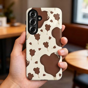 May include: A smartphone case with a cream-coloured background featuring a repeating pattern of brown chocolate-dipped ice cream bars shaped like mouse heads. The case has a black camera module cutout in the upper left corner with three lenses.
