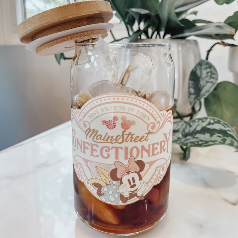 May include: A glass cup with a pink and white label that says "Best Sweets in Town Main Street Confectionery" with a cartoon image of Minnie Mouse. The cup is filled with ice and a dark brown liquid.