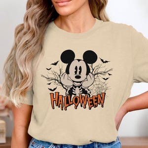 Puede incluir: Una camiseta de color beige con un gráfico de un ratón de dibujos animados con torso de esqueleto, rodeado de siluetas negras de murciélagos, ramas de árboles desnudas y una telaraña. La palabra HALLOWEEN aparece en letras naranjas.