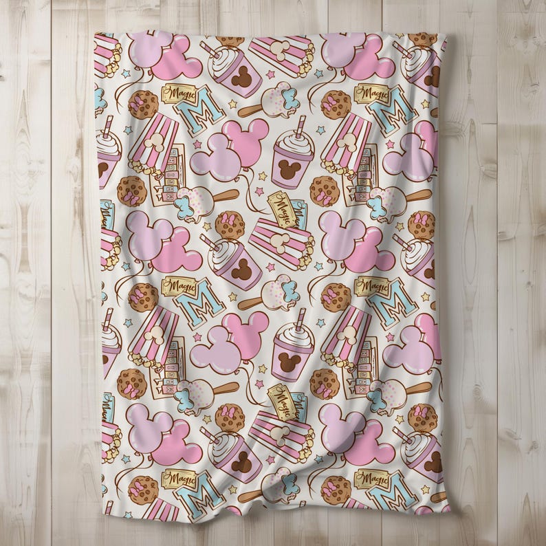 Peut inclure: Un tissu blanc avec un motif rose et marron d'oreilles de Mickey Mouse, de seaux de pop-corn, de coupes de cr&egrave;me glac&eacute;e Mickey Mouse et d'&eacute;toiles. Le texte "Magic" est r&eacute;p&eacute;t&eacute; dans tout le motif.