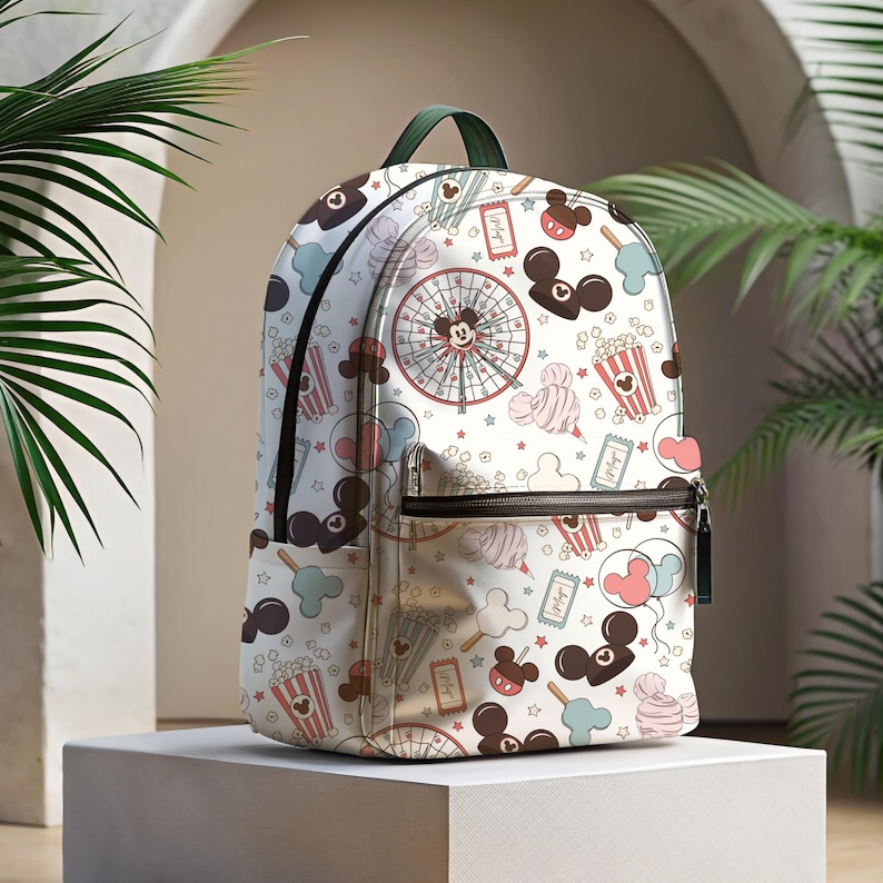May include: White backpack with a colorful print of Mickey Mouse, popcorn, cotton candy, and other carnival-themed imagery. The backpack has a black zipper and a green strap.