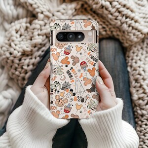 May include: A phone case with a Halloween design featuring Mickey Mouse, ghosts, bats, and the words "Spook Vibes." The case has a beige background with orange, black, and white illustrations. The phone case is held by a person wearing a white sweater.