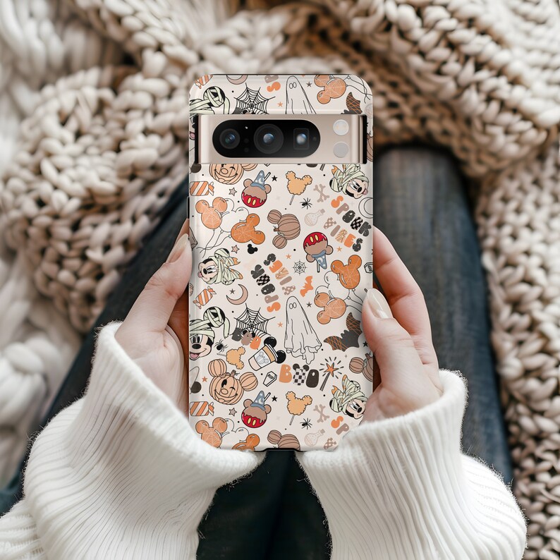 May include: A phone case with a Halloween design featuring Mickey Mouse, ghosts, bats, and the words "Spook Vibes." The case has a beige background with orange, black, and white illustrations. The phone case is held by a person wearing a white sweater.